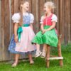 Zwei lachende Mädchen in selbstgenähten Trachtenoutfits nach dem Schnittmuster Kinderdirndl Resli. Die unterschiedlichen Stoffkombinationen zeigen die Vielseitigkeit der Nähanleitung von TOSCAminni für Gr. 80 bis 140.