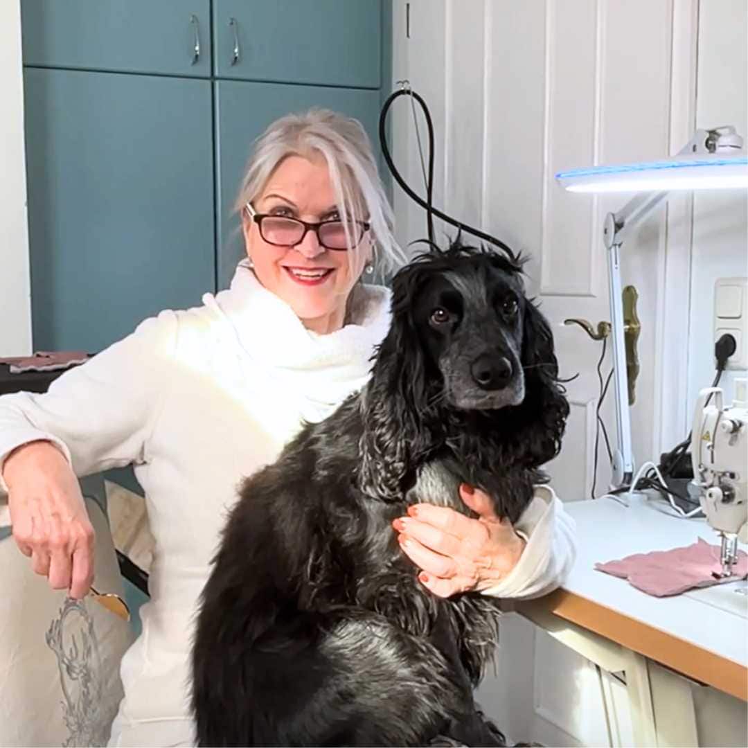 Frau sitzt im Atelier von TOSCAminni-Schnittmanufaktur mit schwarzem Cockerspaniel auf dem Schoß an der Nähmaschine und lacht